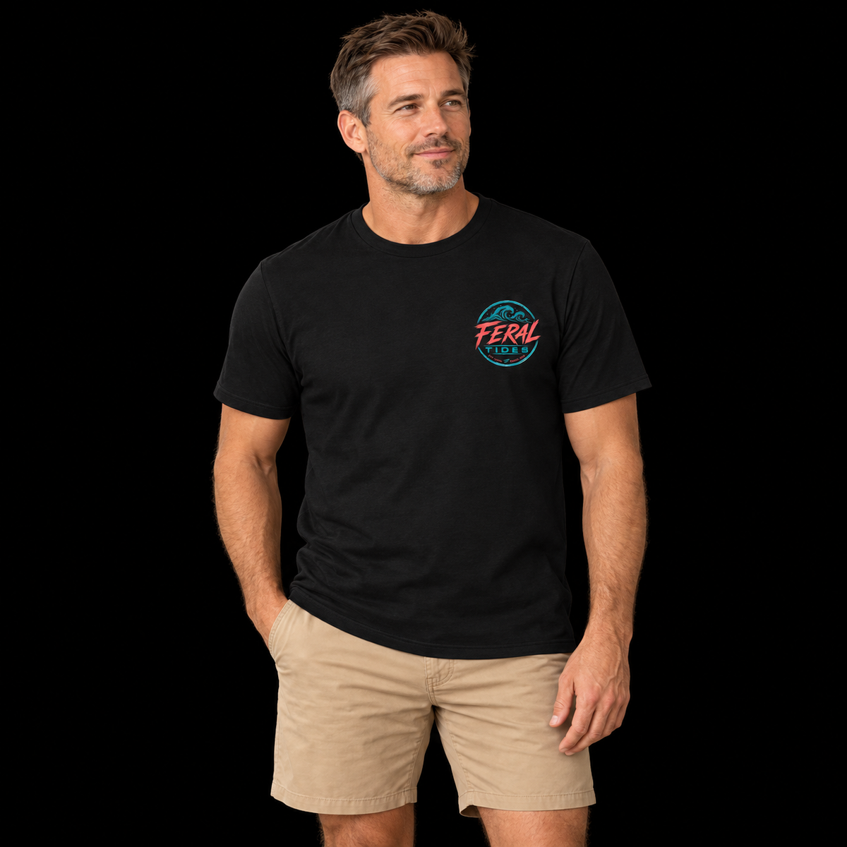 Male model wearing the Black Feral Tides Tidal Rebel Tee, standing with one hand in pocket and looking right, showing the small coral-and-teal wave logo on the front chest against a solid black background.