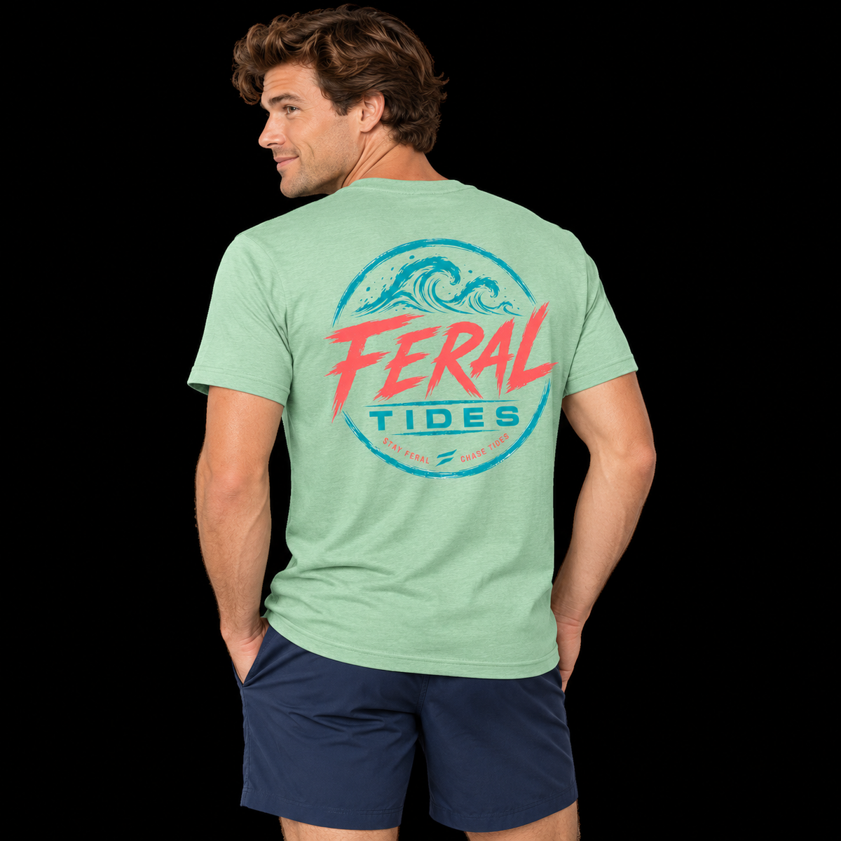 Male model wearing the Heather Neo Mint Feral Tides Tidal Rebel Tee, looking back over left shoulder with hands in pockets, showing the large coral-and-teal wave graphic on the back against a solid black background.