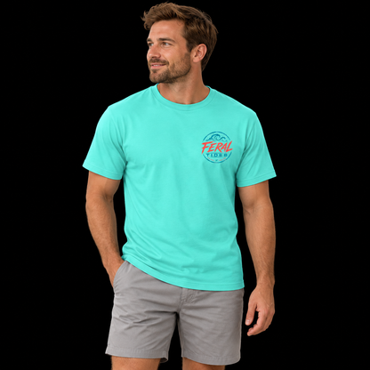 Male model wearing the Mint Feral Tides Tidal Rebel Tee, standing with one hand in pocket and looking left, showing the small coral-and-teal wave logo on the front chest against a solid black background.